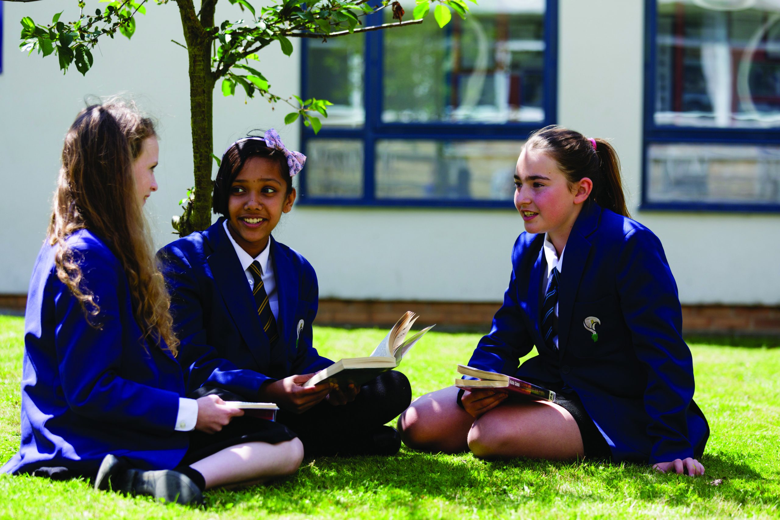 3 girls in school uniform, sitting on the grass, looking at each other and smiling