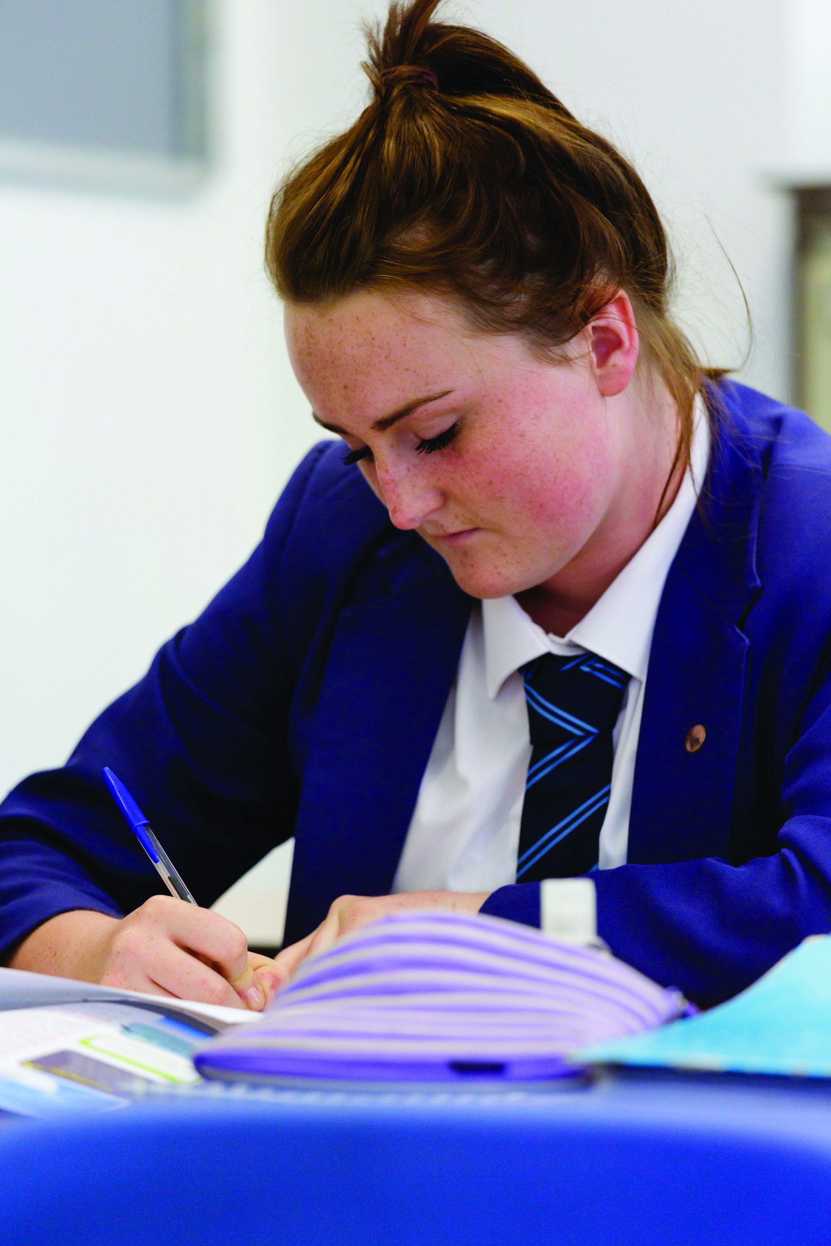 Girl in school uniform sitting at desk, looking down and writing with blue pen