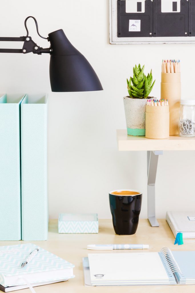 A desk with a notebook and a mug below a shelf with a potted plant and pencils