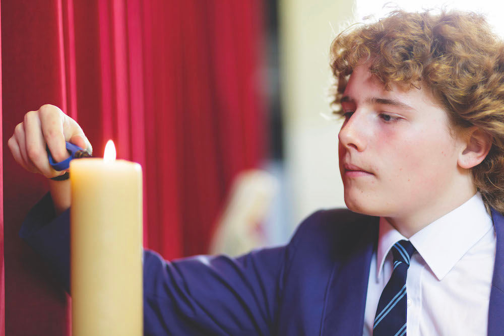 A boy with curly hair lights a yellow candle with a candle lighter in front of a red curtain