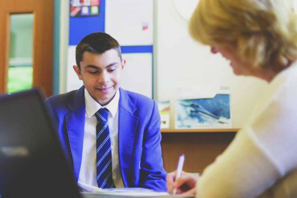 A boy in school uniform sits at a table with a lady, who is smiling and holding a pen