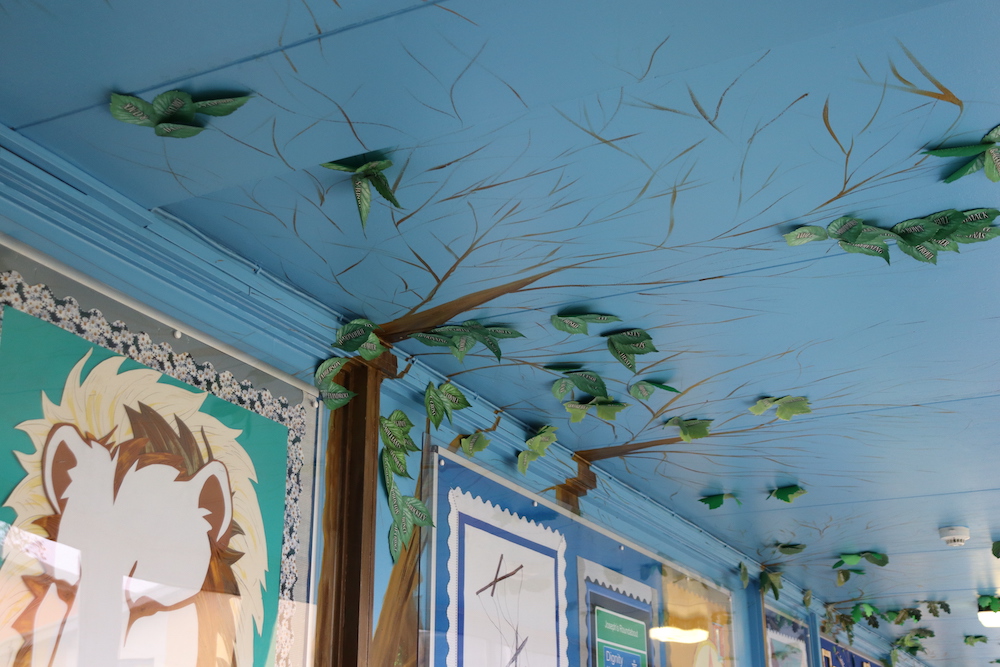 School corridor with posters on the walls, trees painted on the walls and the branches spreading up onto the ceiling, and paper leaves stuck on the branches.